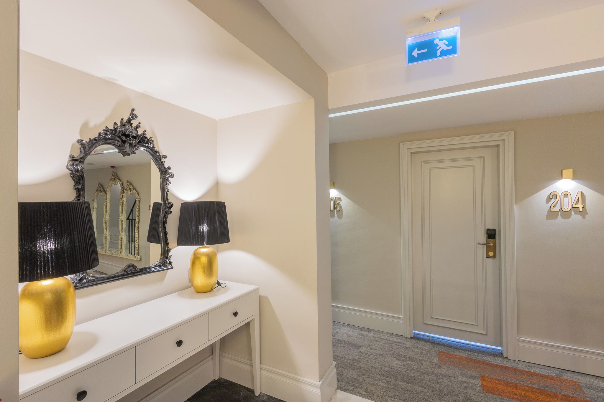 A hotel room with a white console table, a large ornate mirror, two gold lamps, a door with a number sign, and a directional sign above the door.