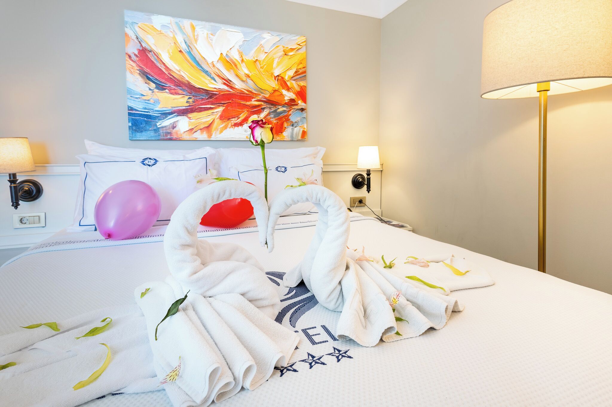 Hotelkamer met een bed, kussens, een wanddecoratie, een roos, handdoeken en een lamp.