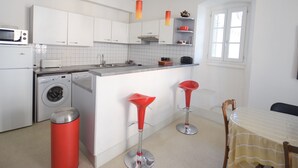 Eine moderne Küche mit weißen Schränken, einer integrierten Waschmaschine, einer Mikrowelle und einem Spülbecken. Zwei rote Barhocker und ein Tisch mit einem gestreiften Tischläufer stehen bereit.