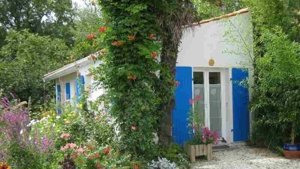 Une maison avec une porte et des fenêtres bleues, entourée d’un jardin où poussent différentes plantes et fleurs.