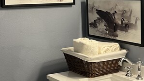 Een badkamer met een witte wastafel, een rieten mand met handdoeken en twee zwart-wit foto's van zwanen.