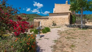 A rustic stone building with a gravel path leading to a fountain, surrounded by vibrant bougainvillea and a clear blue sky.