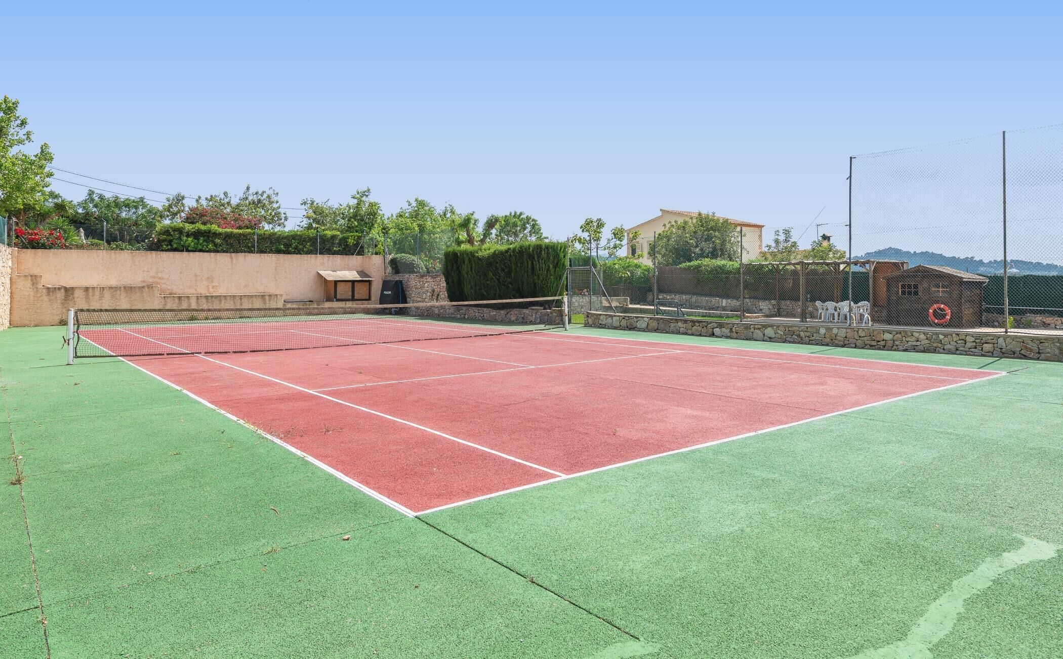 A tennis court with a net, surrounded by a stone wall and greenery.