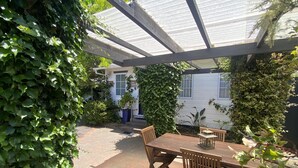 A covered patio area with a wooden table and chairs, surrounded by lush greenery and a white building in the background.