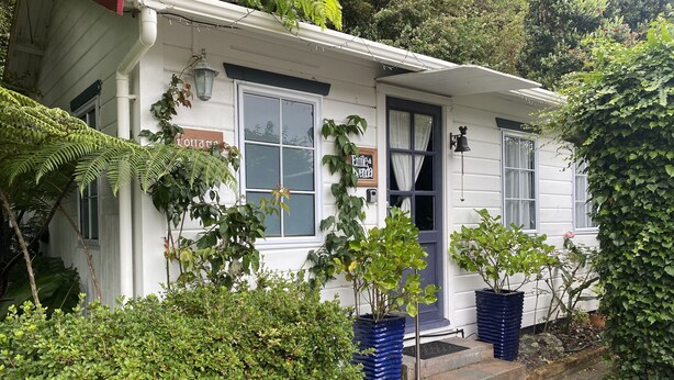 A white building with a blue door and windows, surrounded by greenery and potted plants.