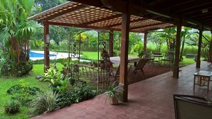 Un espacio al aire libre cubierto con piscina, rodeado de abundante vegetación y un patio con mesas y sillas.