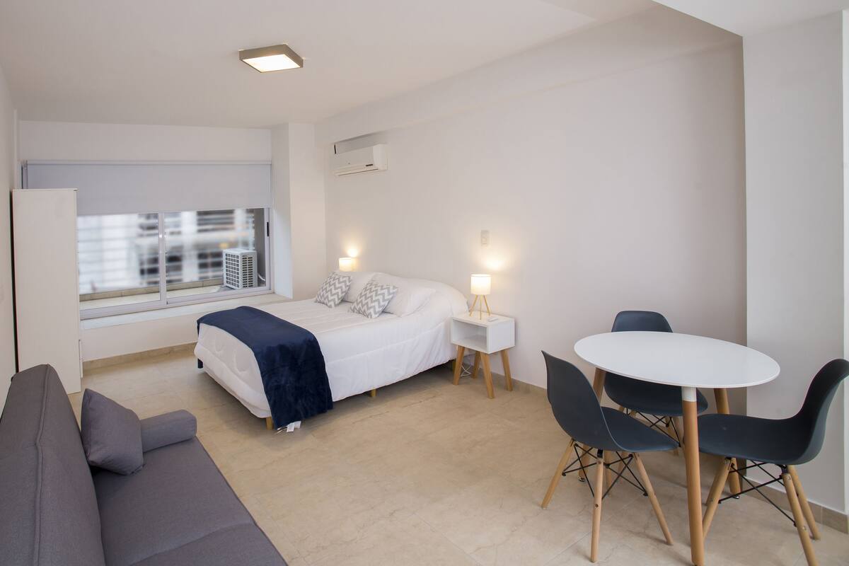 A modern, compact hotel room with a bed, bedside tables, a desk with chairs, a sofa, and a large window offering a city view.