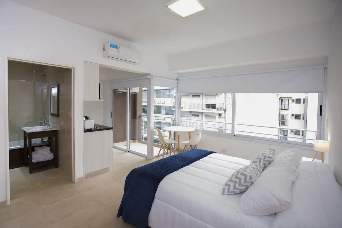 A modern hotel room with a large bed, a bathroom with a sink and mirror, a balcony with a table and chairs, and a city view through the windows.