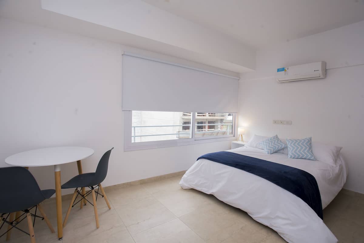 A bedroom with a bed, a window with blinds, a table with chairs, and an air conditioning unit on the wall.