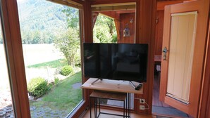 A room with a large flat-screen TV on a wooden stand, a door with a window, and a view of a green landscape. - "schossbach" Baubiologische Ferienwohnung 60 qm, Zwei Extra Schlafzimmer (Aschau im Chiemgau)