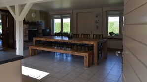 A spacious dining area with a large wooden table and benches, a brick fireplace, and large windows with a view of greenery. - holiday home for 10 people (Oostvleteren)