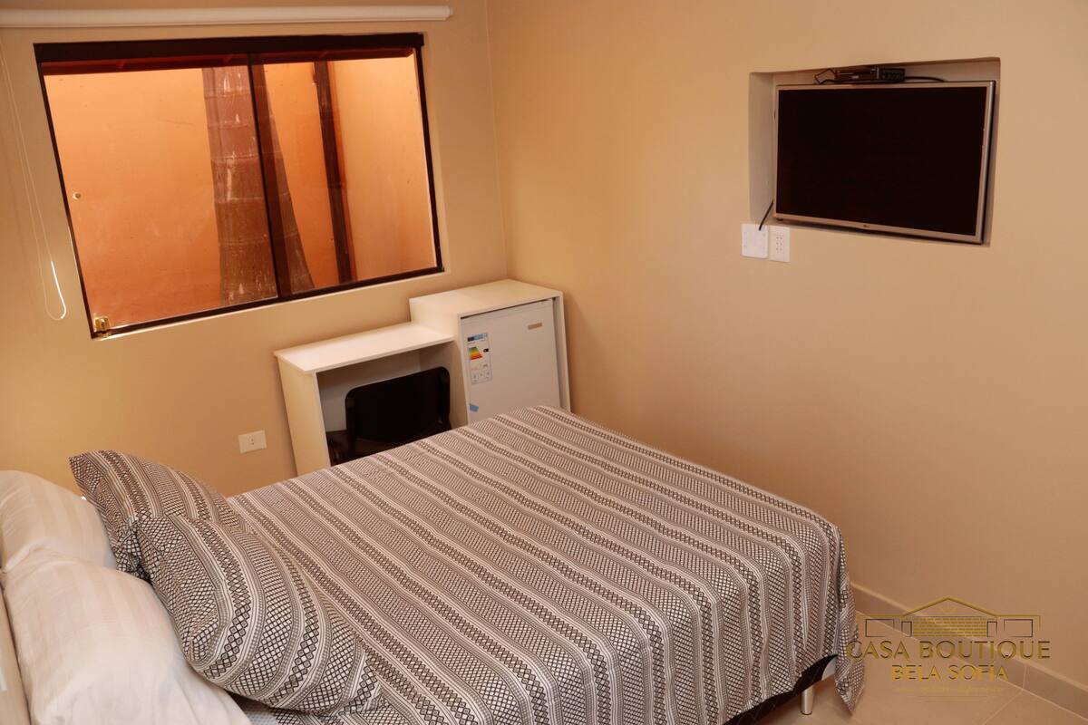 A bedroom with a bed, a window, a wall-mounted TV, and a small refrigerator.