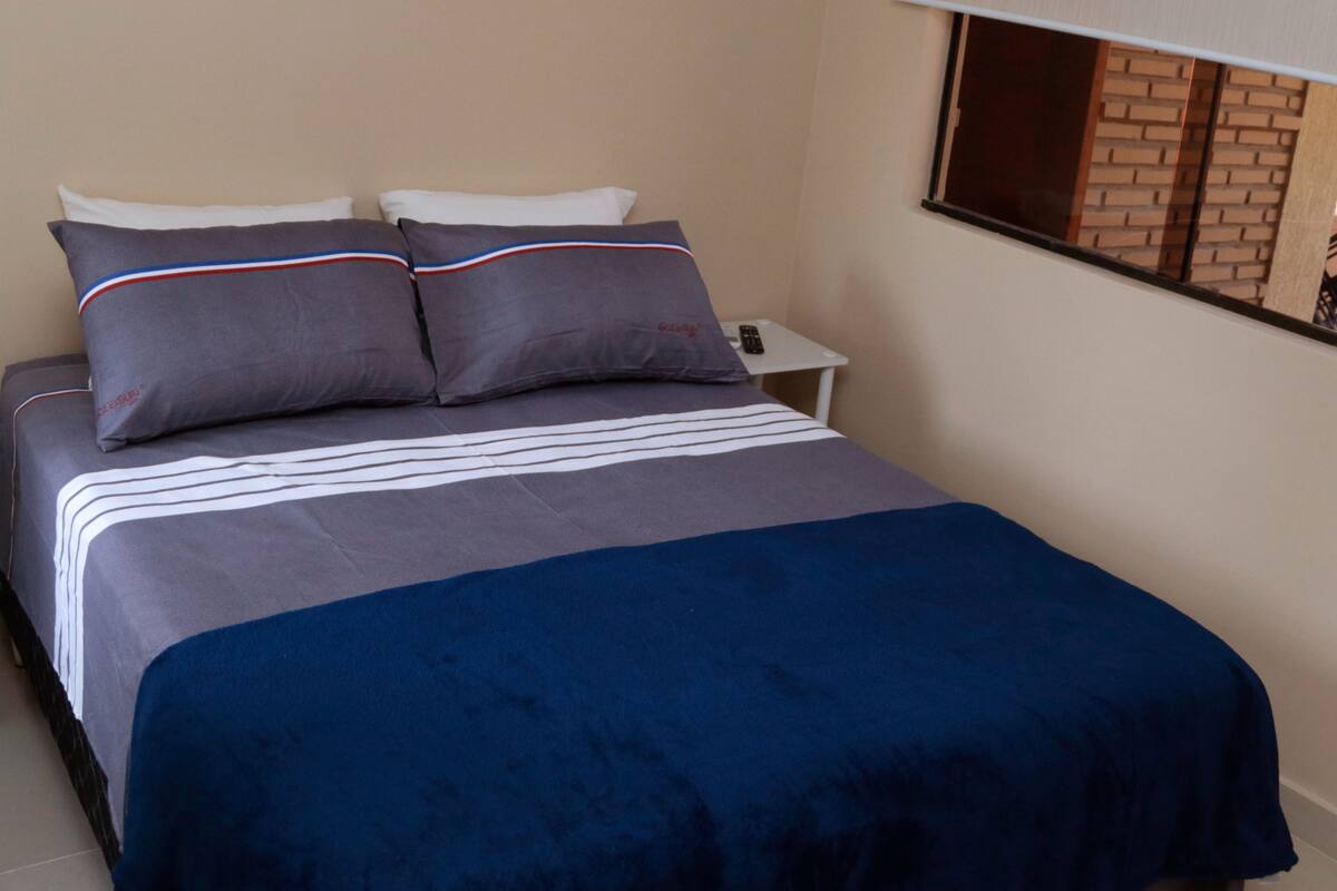A bed with a grey and blue bedspread, two pillows with red and white stripes, and a white nightstand.