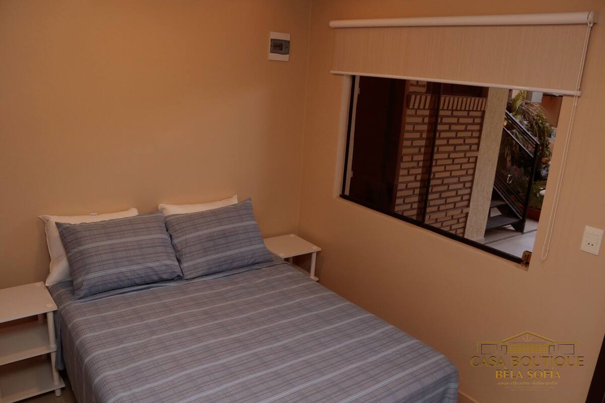A bedroom with a bed, two bedside tables, a window with a view of a brick wall, and a wall-mounted light switch.