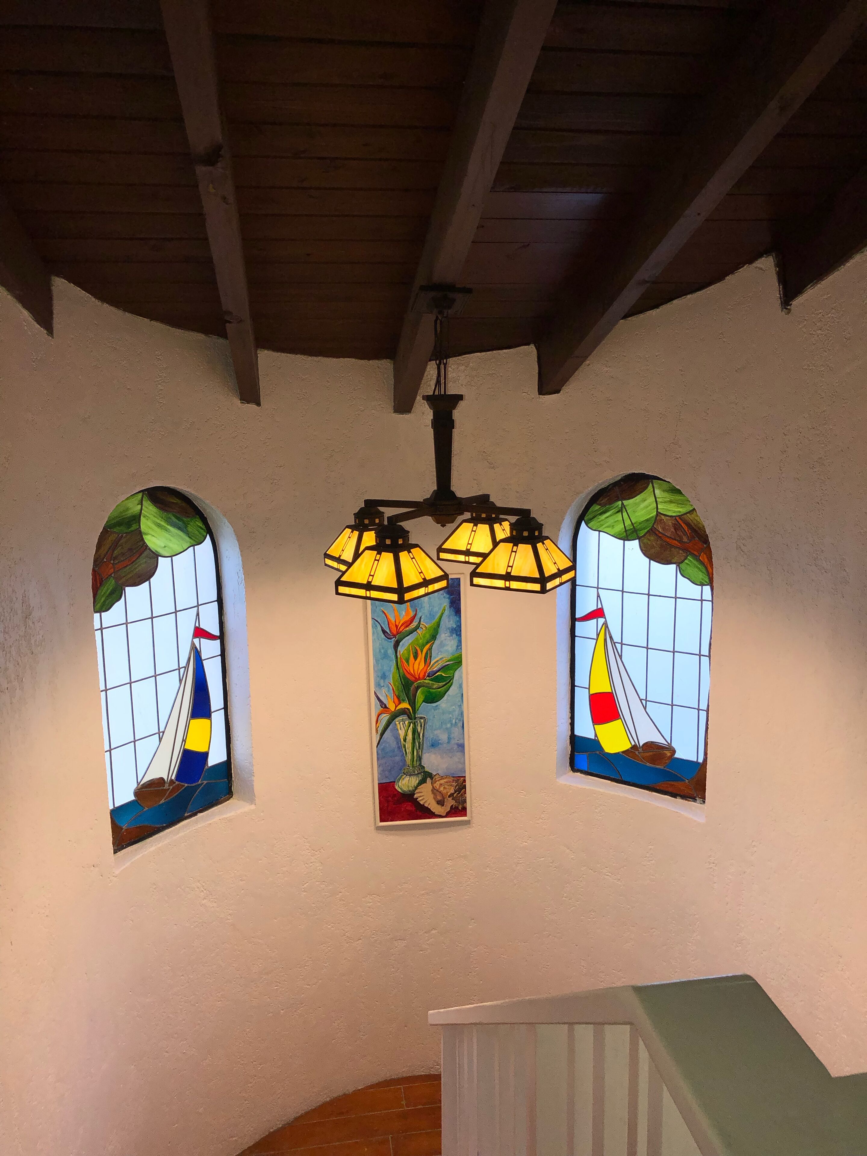A room with three stained glass windows depicting sailboats and a vase with flowers, a wooden ceiling, and a white bench.