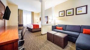A hotel room with a wooden desk, a grey sofa, a bed with a red blanket, a small table, and three framed pictures on the wall.