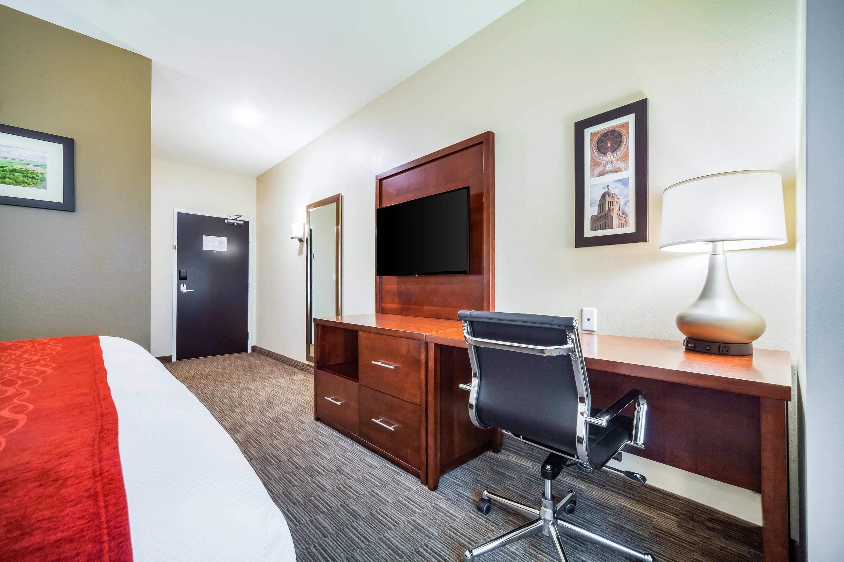 A hotel room with a bed, a desk with a chair, a TV, a lamp, and a door.