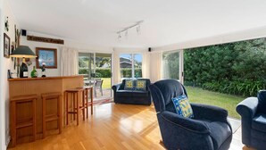 A spacious living area with a wooden floor, a dark blue armchair with a cushion, a sofa, and a bar area with stools. - Matapouri Beach Family Haven - Matapouri Home (Matapouri)