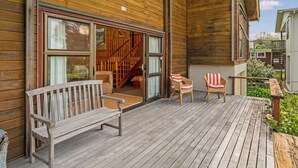 Un terrazzo in legno con una panchina e due sedie in vimini, una scala in legno che conduce a un livello superiore e un piccolo giardino.