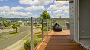 A modern house with a wooden deck, a glass door, and a view of a road and greenery.