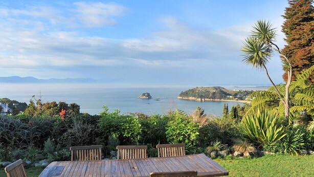 A wooden table and chairs set outdoors with a view of the sea and hills in the background.