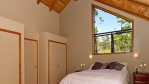 A bedroom with a large window, a bed with white bedding, two bedside tables, and a wooden ceiling.