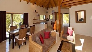 A spacious living area with a high wooden ceiling, a dining table with chairs, a sofa with cushions, and a kitchen area with a counter and shelves.
