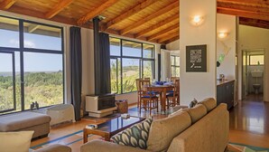 A spacious living area with a wooden ceiling, large windows offering a view of the landscape, a fireplace, and a dining area. - Hahei Hideaway - Hahei Holiday Home (Hahei)