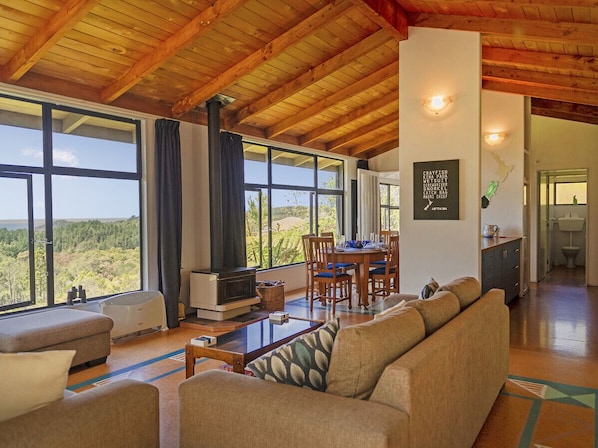 A spacious living area with a wooden ceiling, large windows offering a view of the landscape, a fireplace, and a dining area.