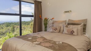 A bedroom with a large bed, a view of the outdoors, and a nightstand with a lamp. - Hahei Hideaway - Hahei Holiday Home (Hahei)