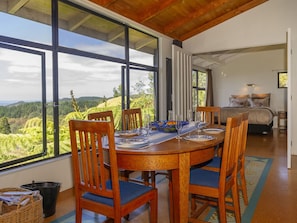 A dining area with a table set for four, large windows with a view of greenery, and a bed in the background.