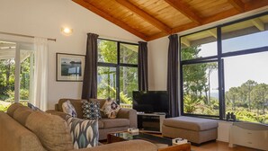 A spacious living room with a large TV, comfortable seating, and large windows offering a view of the outdoors. - Hahei Hideaway - Hahei Holiday Home (Hahei)