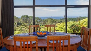A round wooden table with four chairs, a bowl of oranges, and a view of a lush landscape through large windows. - Hahei Hideaway - Hahei Holiday Home (Hahei)