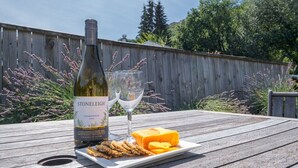 A bottle of Stoneleigh Chardonnay 2018, two wine glasses, a plate with cheese and crackers, and a wooden table outdoors.