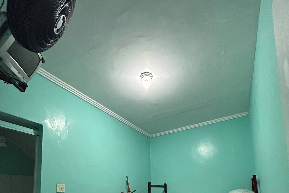 Una habitación pequeña con cama individual, paredes color turquesa, ventilador de techo y toma de corriente visible.