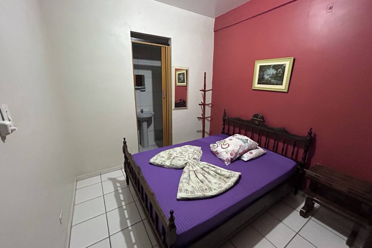 Un dormitorio con cama, cabecero de madera, pared roja, un cuadro enmarcado y una puerta blanca que da al baño.