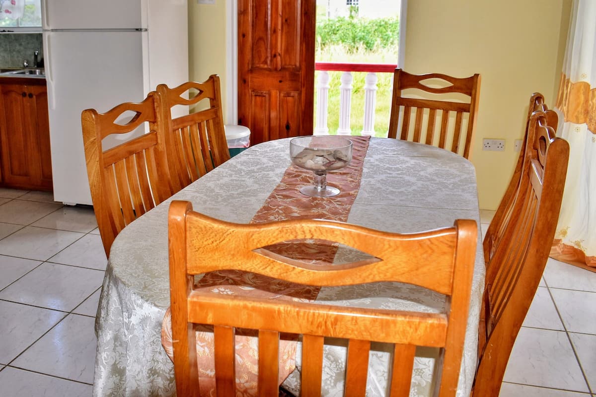 Une table à manger recouverte d’une nappe en marbre, entourée de chaises en bois dont les dossiers sont sculptés. Un verre d’eau est posé sur la table.