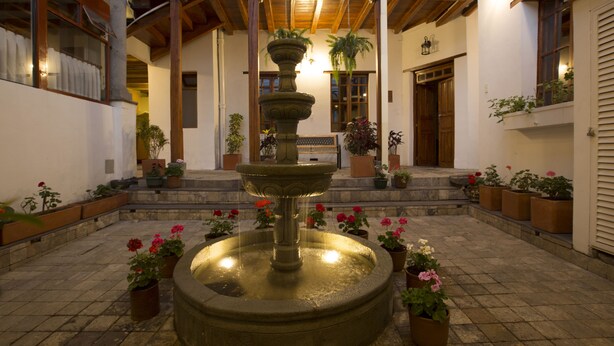Um pátio com uma fonte central, rodeado por vasos de plantas e uma escadaria de madeira que dá acesso a um andar superior.