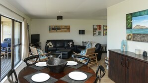 Et spiseområde med et rundt bord, stole og en skål i midten. Der er en stue med en sofa og et fjernsyn i baggrunden.