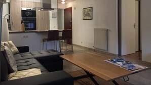 En kompakt opholdsstue med sofa, spisebord og et tekøkken med skabe og hårde hvidevarer.