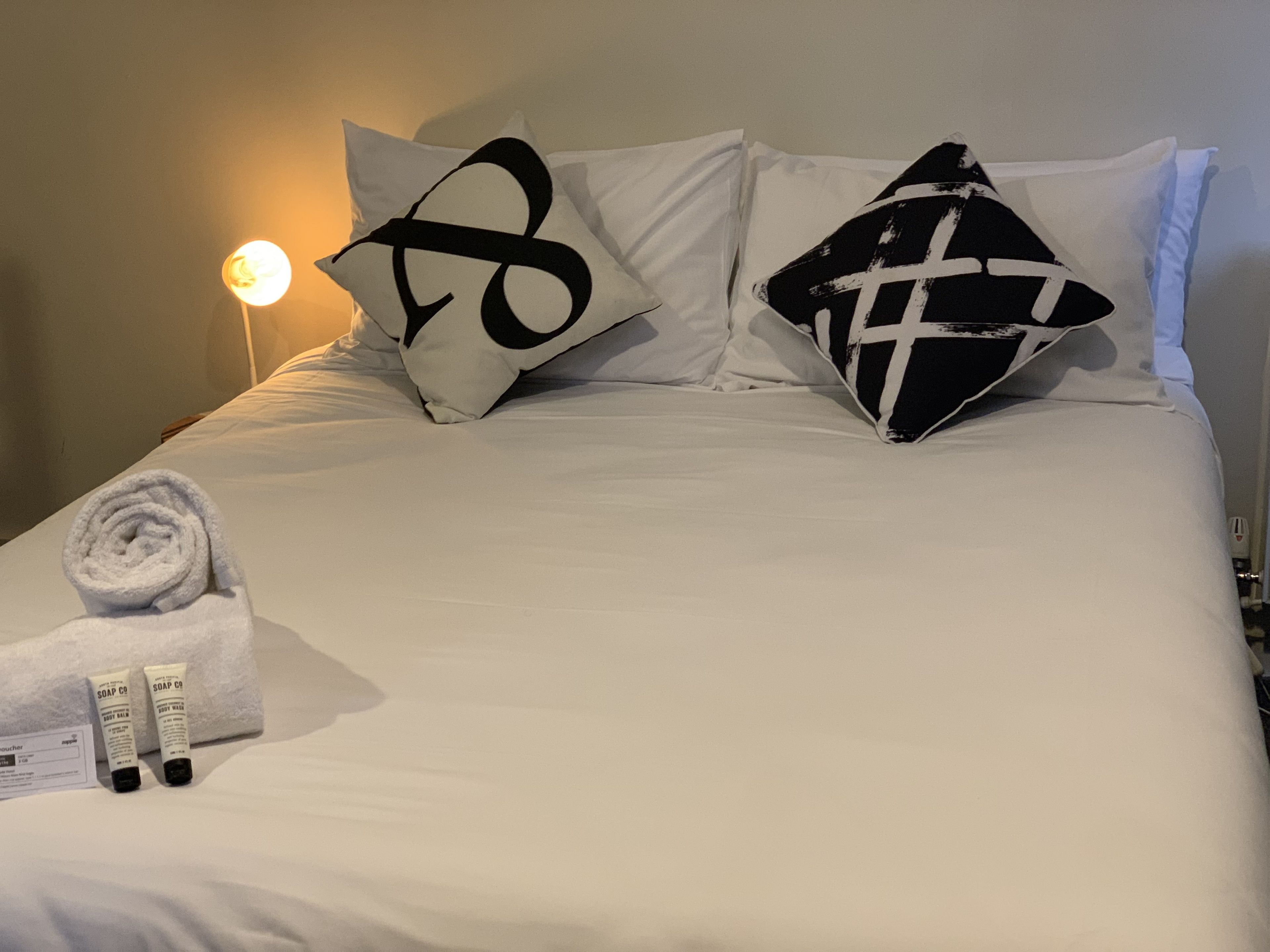 Una cama bien hecha con ropa blanca y dos almohadillas decorativas, una con diseño blanco y negro. Un toalla blanca enrollada y dos pequeñas botellas de jabón están sobre la cama.