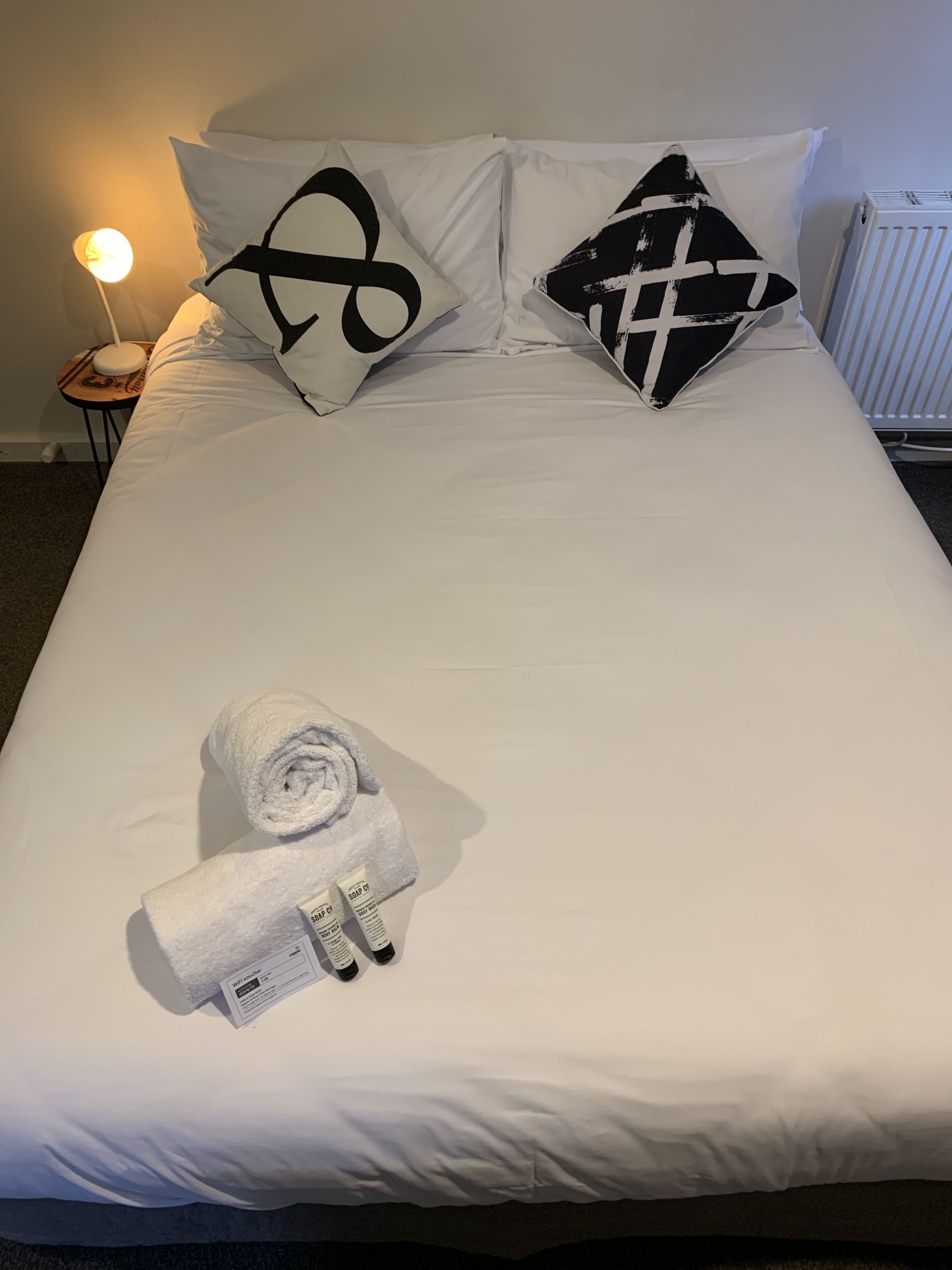 Ein ordentlich bezogenes Bett mit weißer Bettwäsche, zwei schwarz-weißen Kissen, einem aufgerollten weißen Handtuch und einer Seifenflasche.