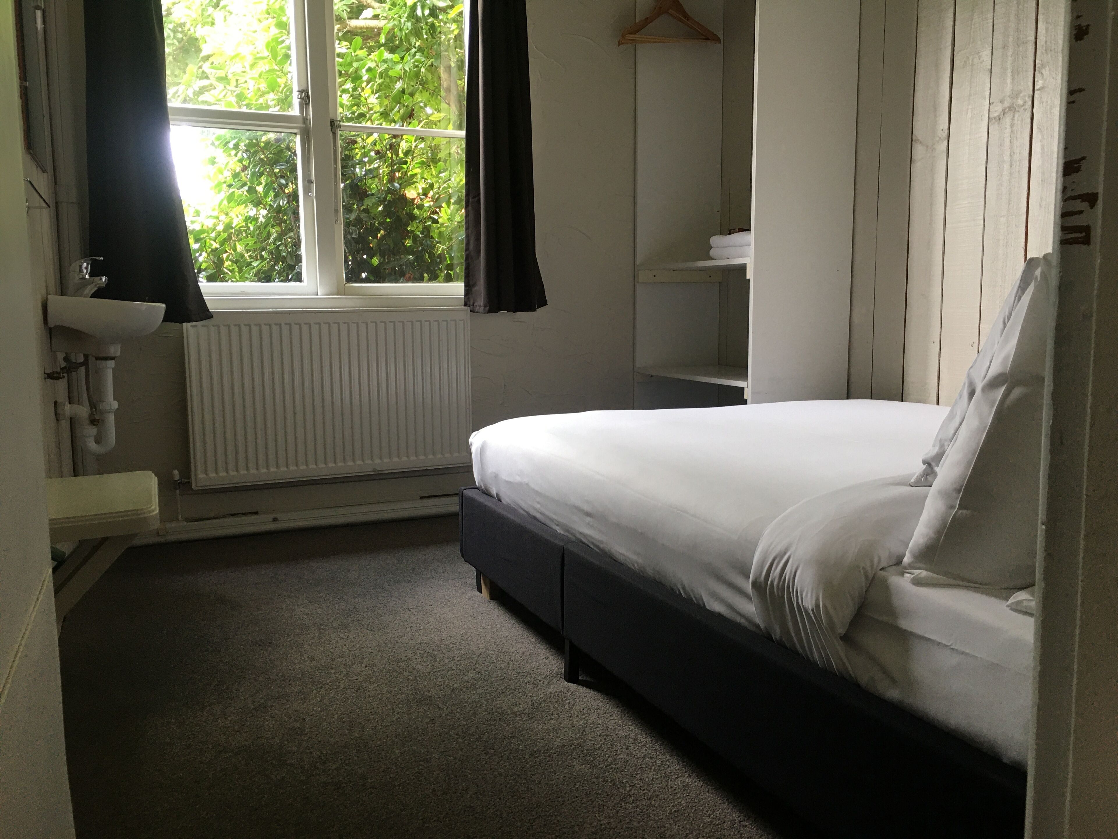 A bedroom with a bed, a window with curtains, a radiator, a shelf, and a hanging clothes rack.