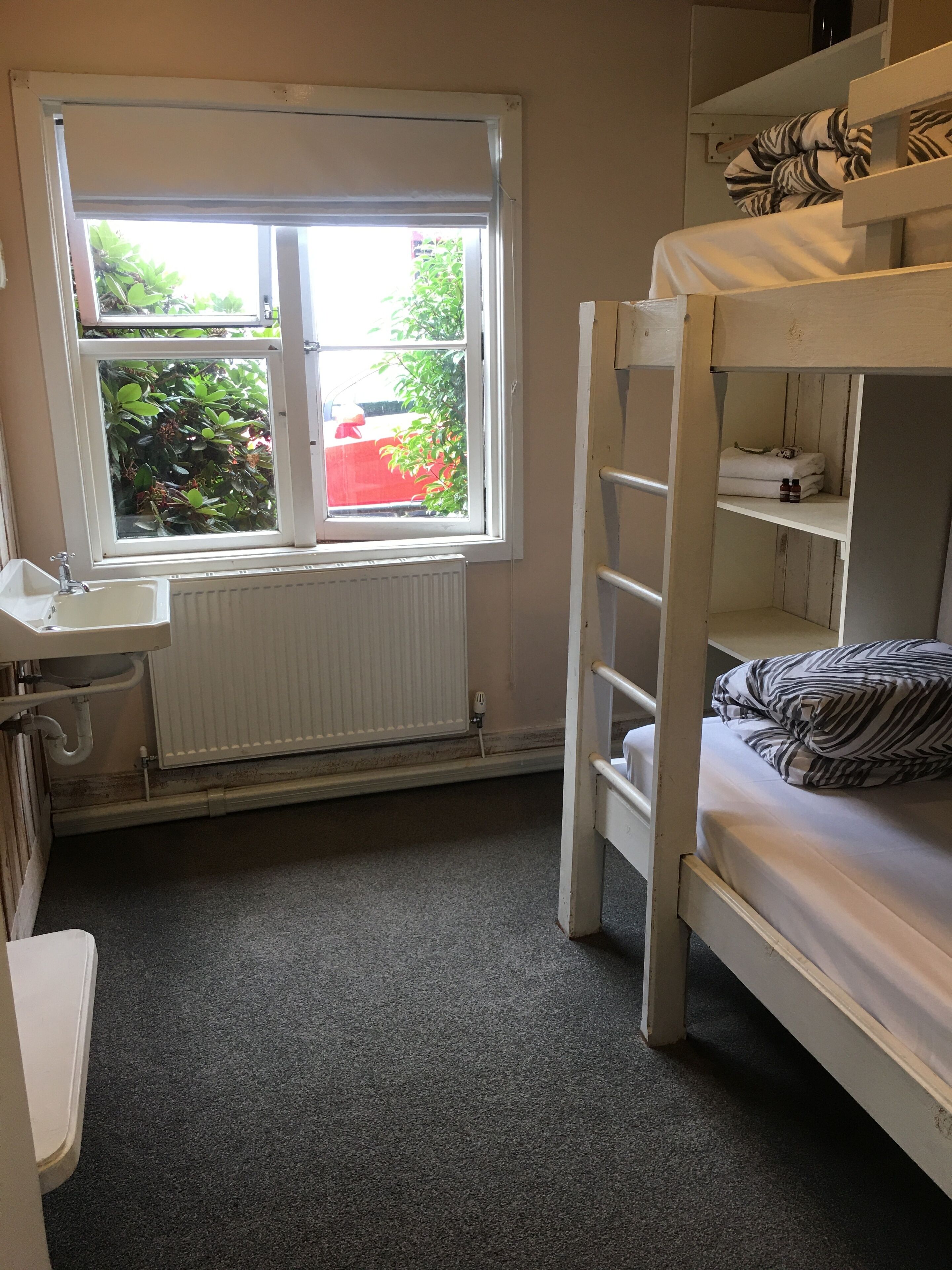 Una cama litera con escalera, una ventana con vistas a follaje, un radiador y un lavamanos pequeño y blanco.