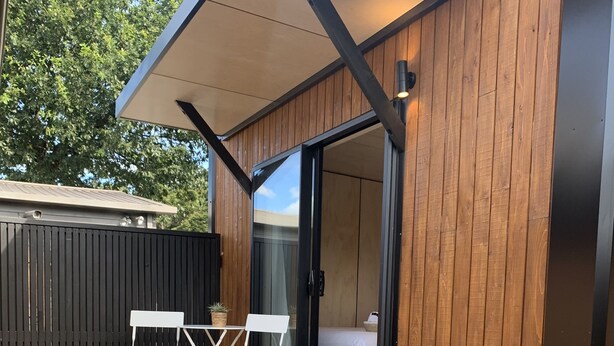A wooden deck with a small table and chairs, a bed in the background, and a set of wooden steps leading to a door. - Snowman Lodge (Ohakune)