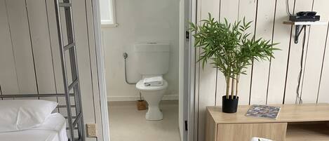Habitación de hotel con literas, un inodoro, una planta en maceta, un televisor y una mesita de noche con toalla y artículos de aseo.