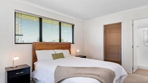 Un dormitorio con cabecera de madera, cama con ropa de cama blanca, mesita de noche con lámpara, ventana con persianas y vestidor.