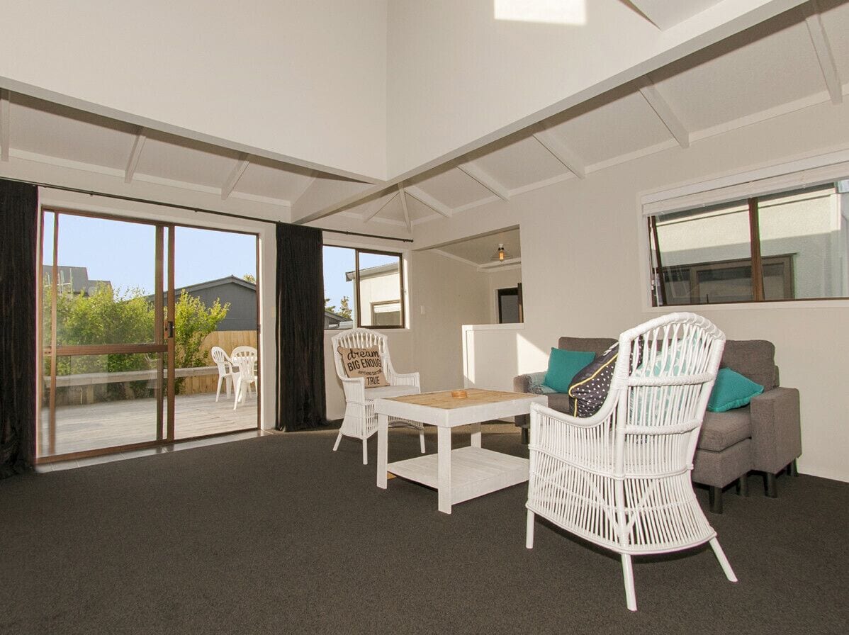 Una habitación amplia con un gran ventanal corredera que da a un espacio exterior, una silla de mimbre blanca, un sofá gris y una mesita blanca.