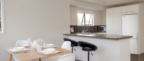 A modern kitchen with white cabinetry, a central island with bar stools, a dining table set for four, and a large window providing natural light.