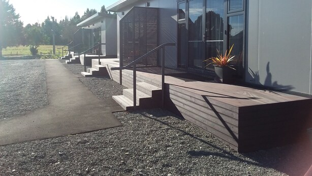 A modern building with a staircase leading to a covered entrance, a paved driveway, and a potted plant near the entrance. - Pine Lake View Lodge (Kaiapoi)